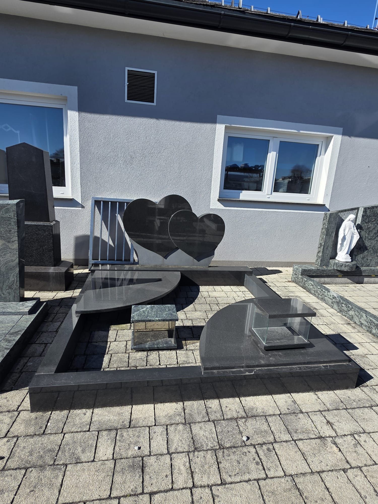 Terrasse mit schwarzer, herzförmiger Bank und passendem Betontisch auf grauen Pflastersteinen neben einem Haus