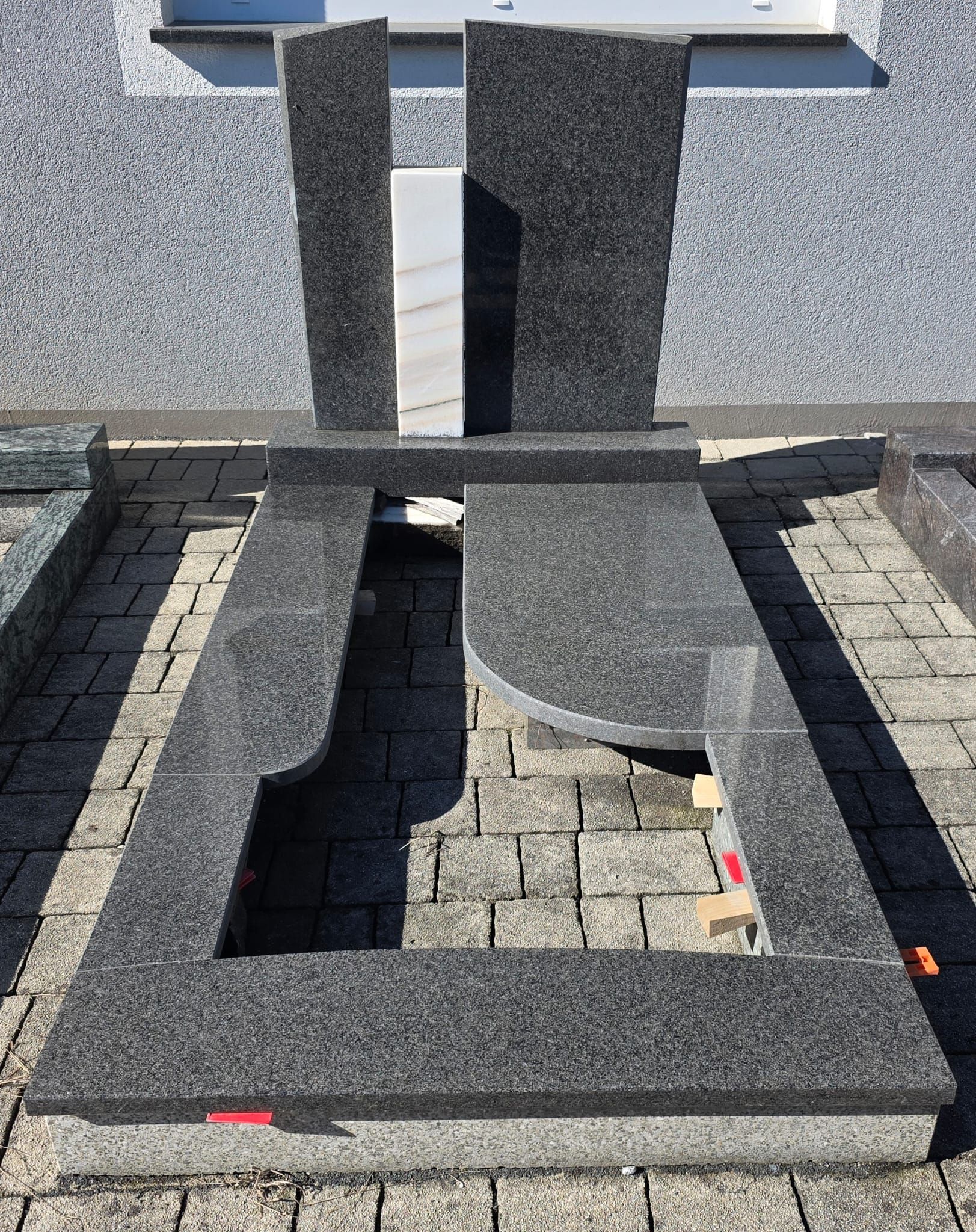 Schwarzes, steinumrahmtes Grabfeld mit rechteckiger Öffnung und einem stehenden Grabstein auf Kies.