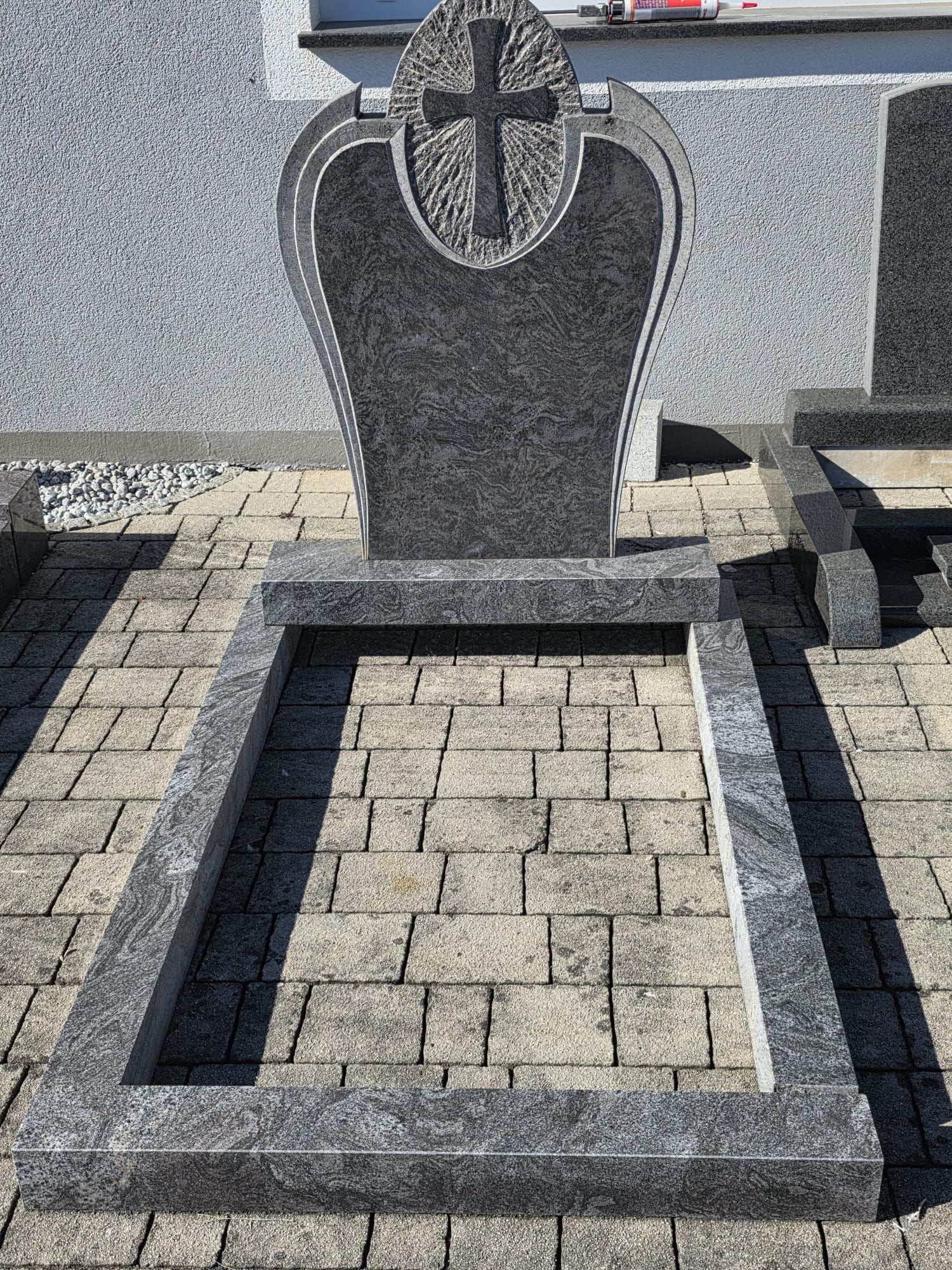 Granitgrabstein mit Kreuz auf einem gepflasterten Friedhofsplatz, von vorne gesehen.