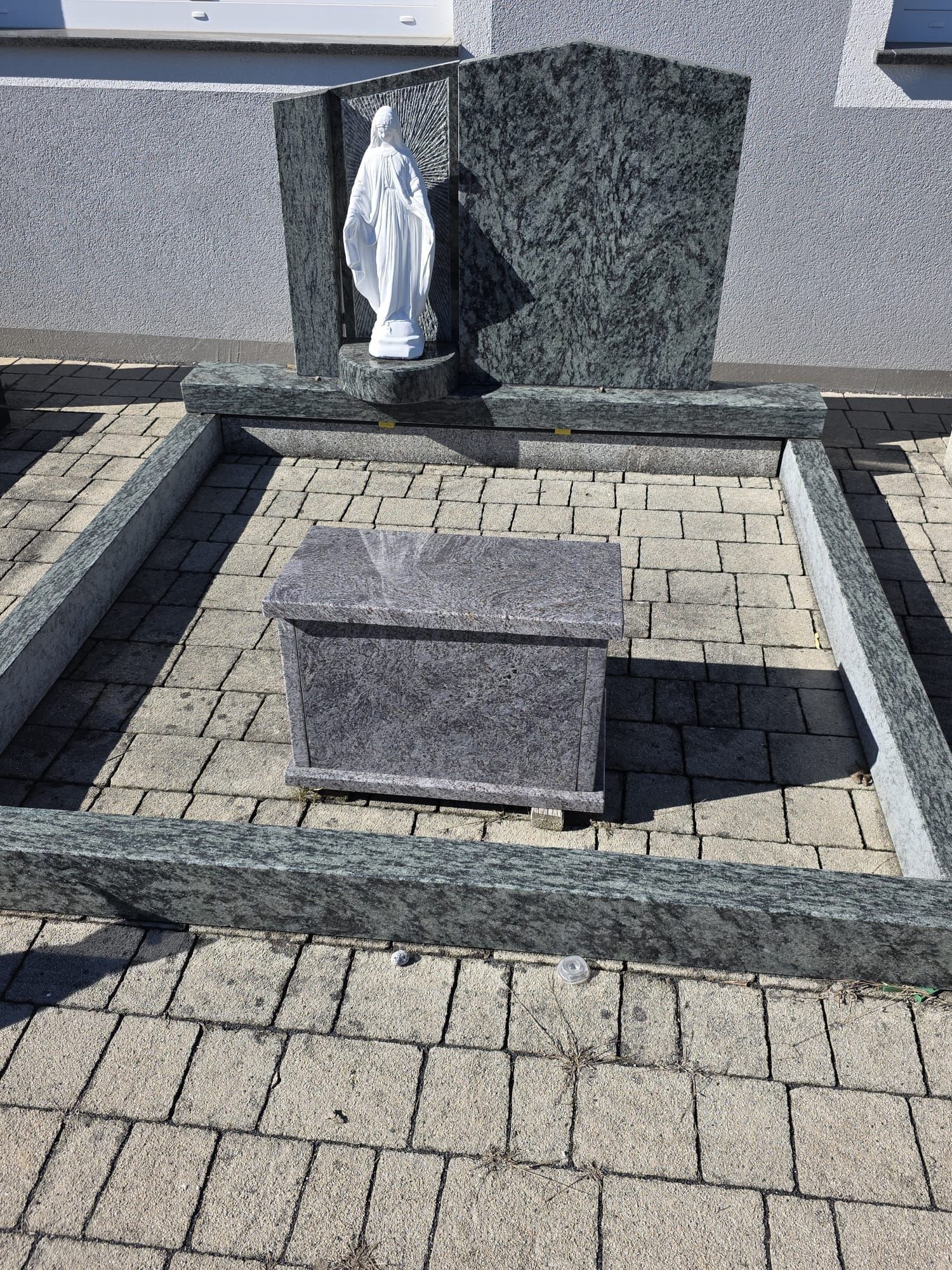 Kleiner Steinschrein mit einer Statue und einem zentralen rechteckigen Sockel auf einem gepflasterten Gedenkhof