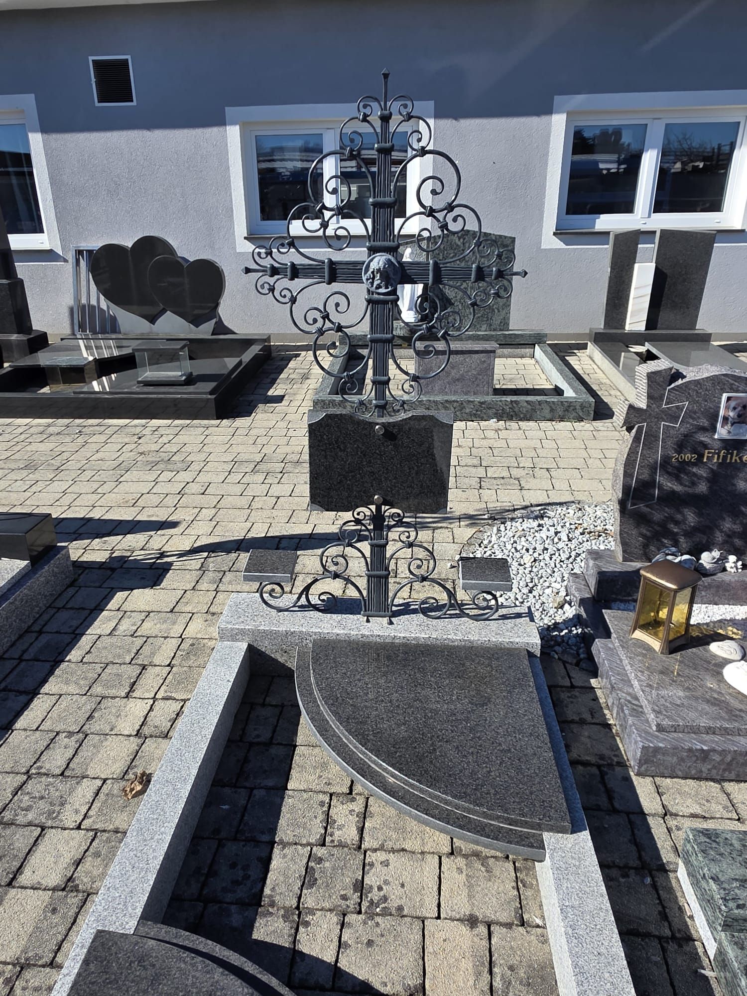 Friedhof mit kunstvollem schwarzem Metallkreuz und herzförmigen Grabsteinen neben einem grauen Gebäude