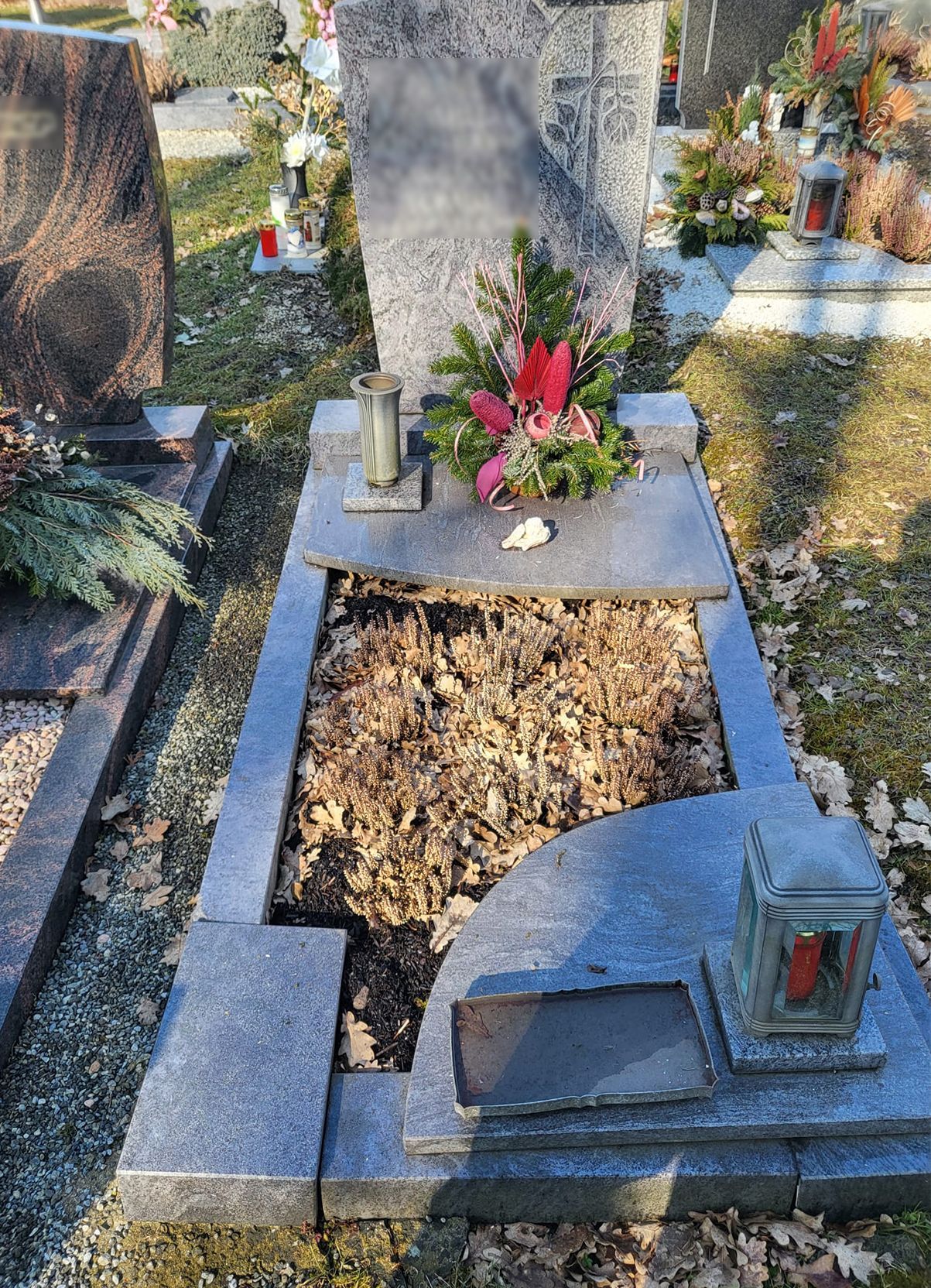 Grabstein mit Blumen und ein offenes Grab auf einem Friedhof, umgeben von grauen Steinmarkierungen.