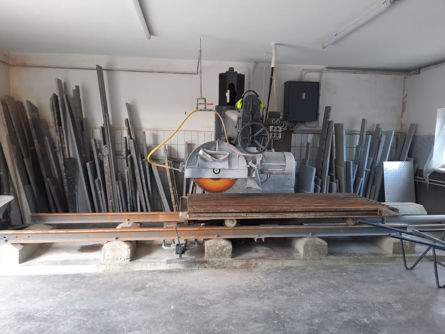 Werkstatt mit einer Metallsäge auf einer Werkbank, umgeben von gestapelten Stahlstangen und -brettern.