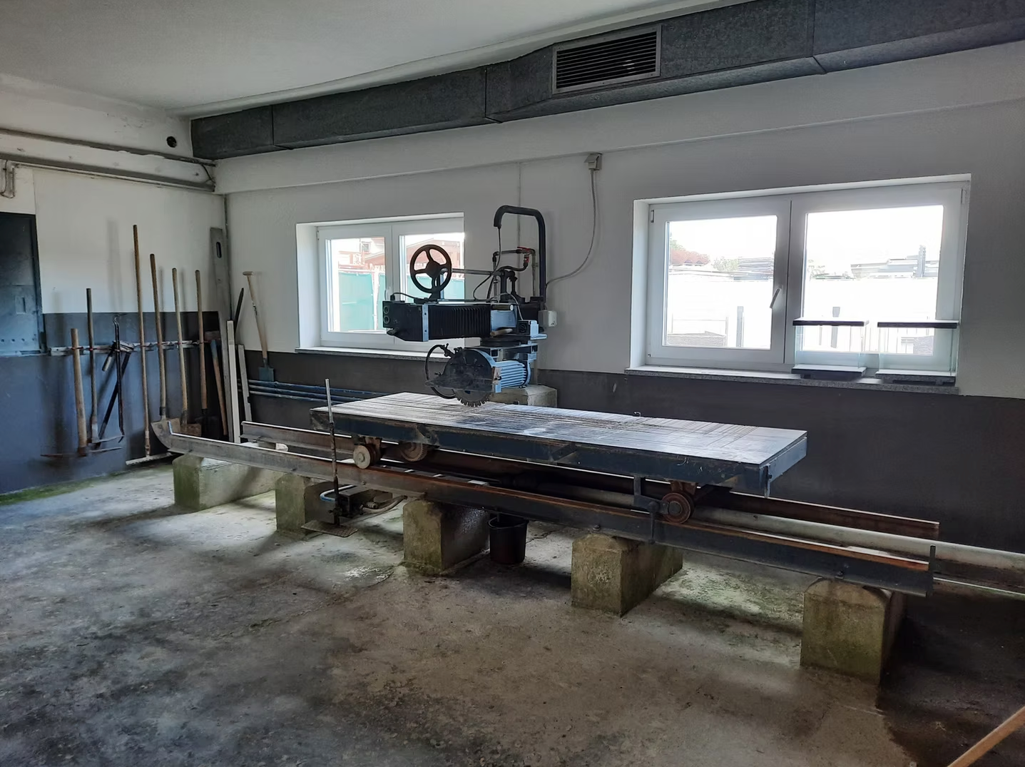 Industriehalle mit einer großen Metallmaschine und Werkbank unter hellen Fenstern, Werkzeuge entlang der Wand aufbewahrt.