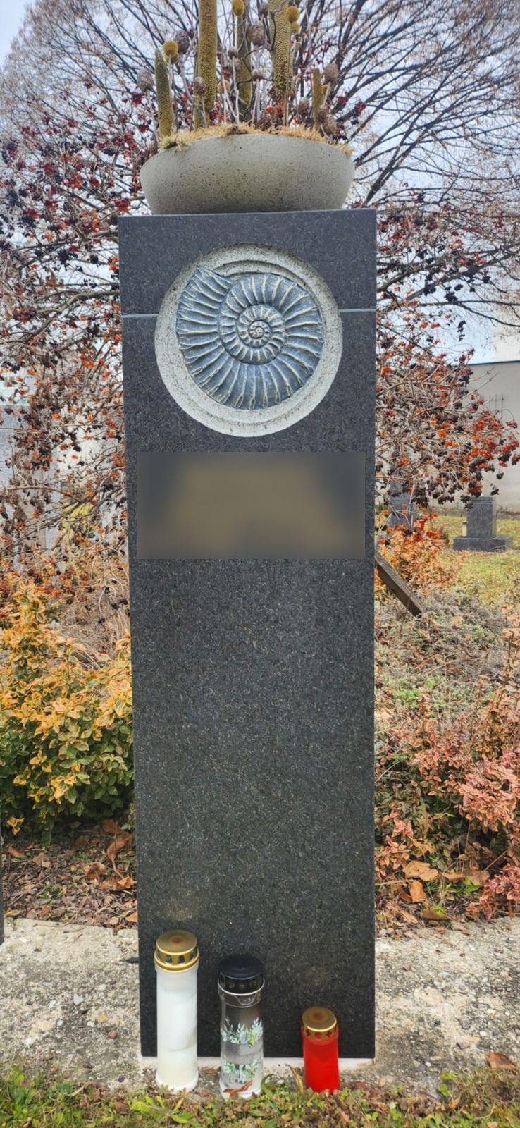 Hohes Granitdenkmal mit kreisförmigem Emblem, im Freien zwischen Herbstbäumen aufgestellt, mit Kerzen am Sockel