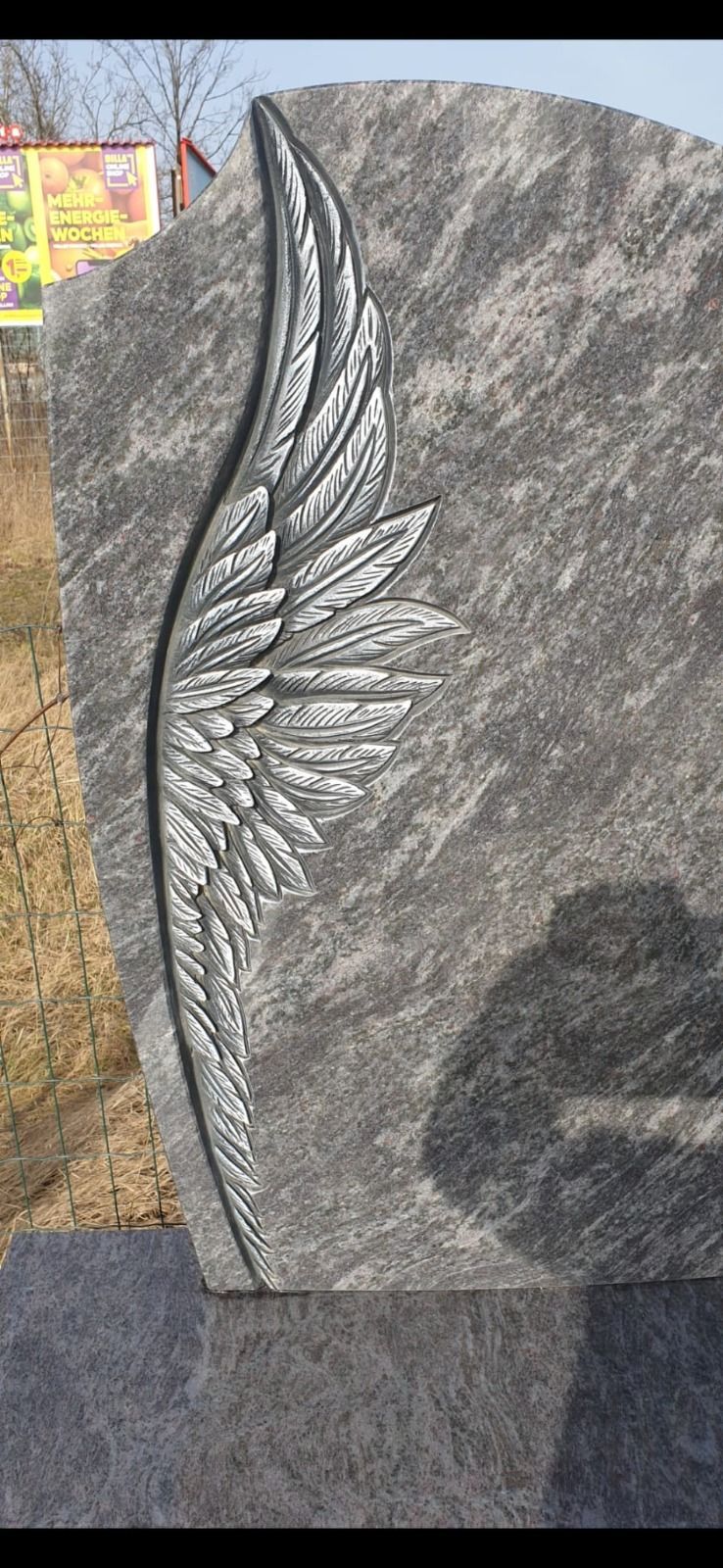 Gedenkstein aus grauem Stein mit eingraviertem Engelsflügel und Federdetails im Freien