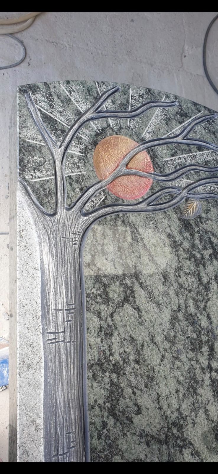 Steinrelief eines Baumes mit Ästen über einer kreisförmigen Sonne und einer strukturierten dunklen Tafel