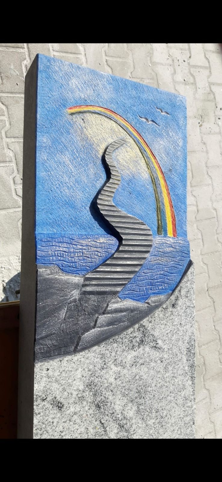 Abstraktes Mosaik eines gewundenen schwarzen Pfades über blauem Wasser, im Hintergrund spannt sich ein Regenbogen.