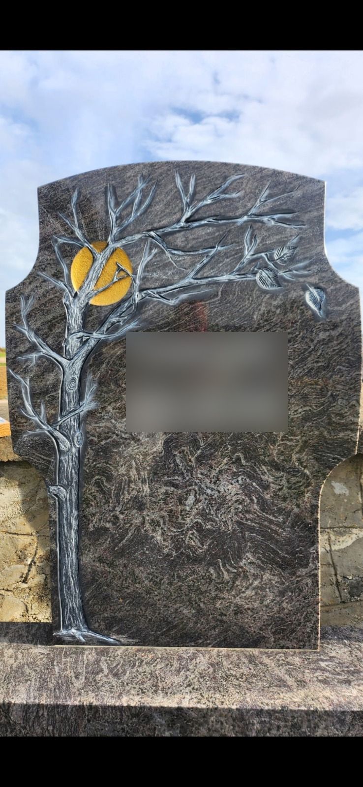 Dunkelgrauer Gedenkstein mit einem weißblättrigen Baum und gelben herzförmigen Blättern unter einem bewölkten Himmel