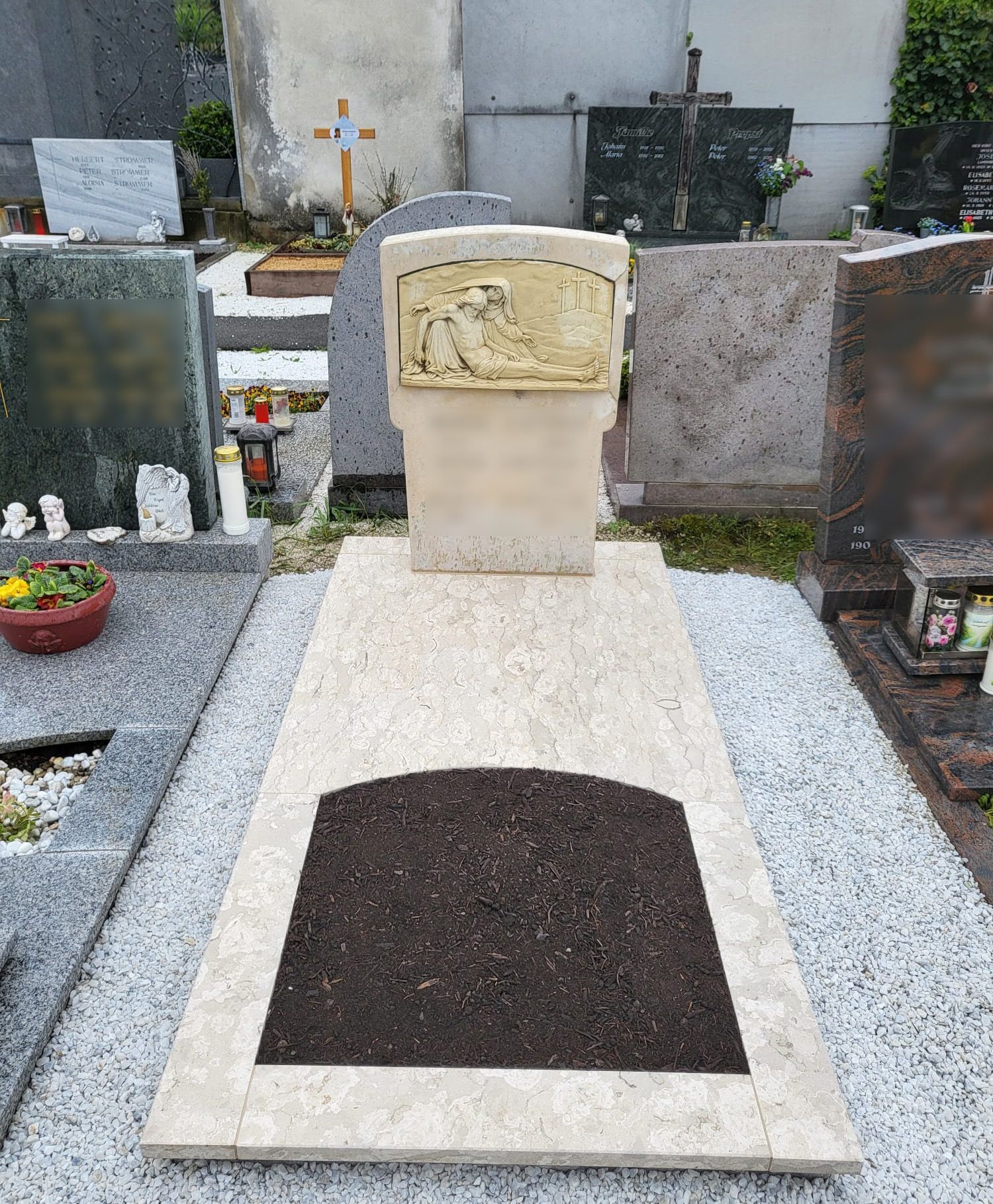 Grabstein mit beiger Gedenktafel und dunklem Blumenbeet auf einem Friedhof, umgeben von anderen Denkmälern.