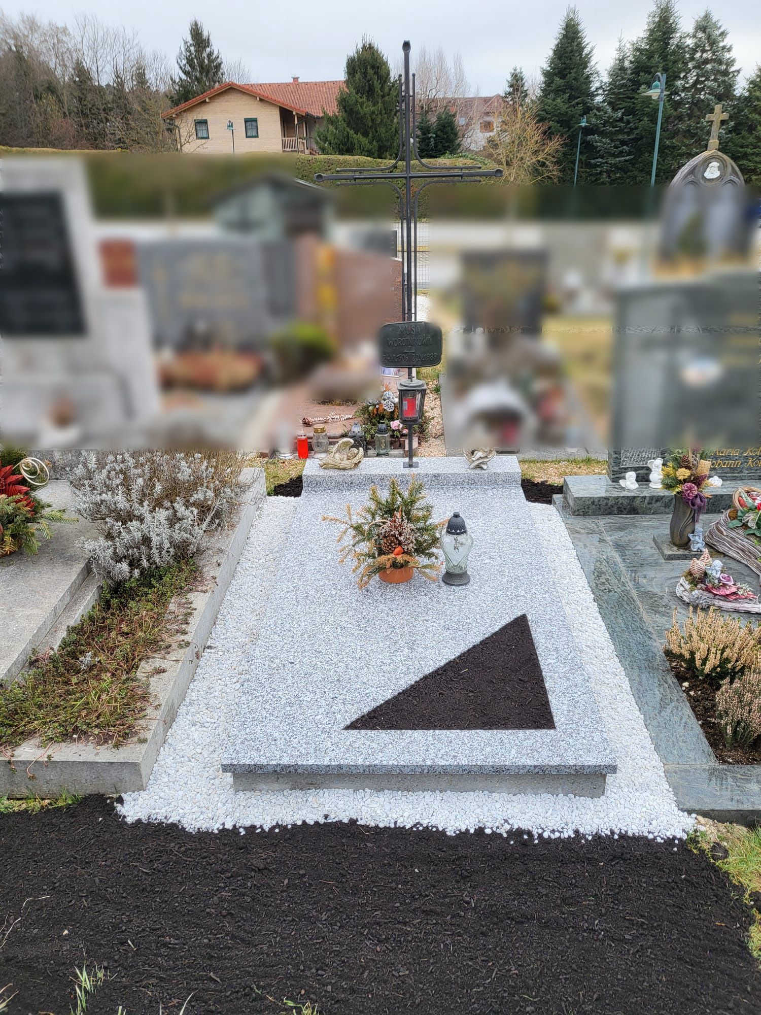 Grabstein mit weißem Kies und einem schwarzen Dreieck auf einem Friedhof, mit Blumen und verschwommenen Grabsteinen.