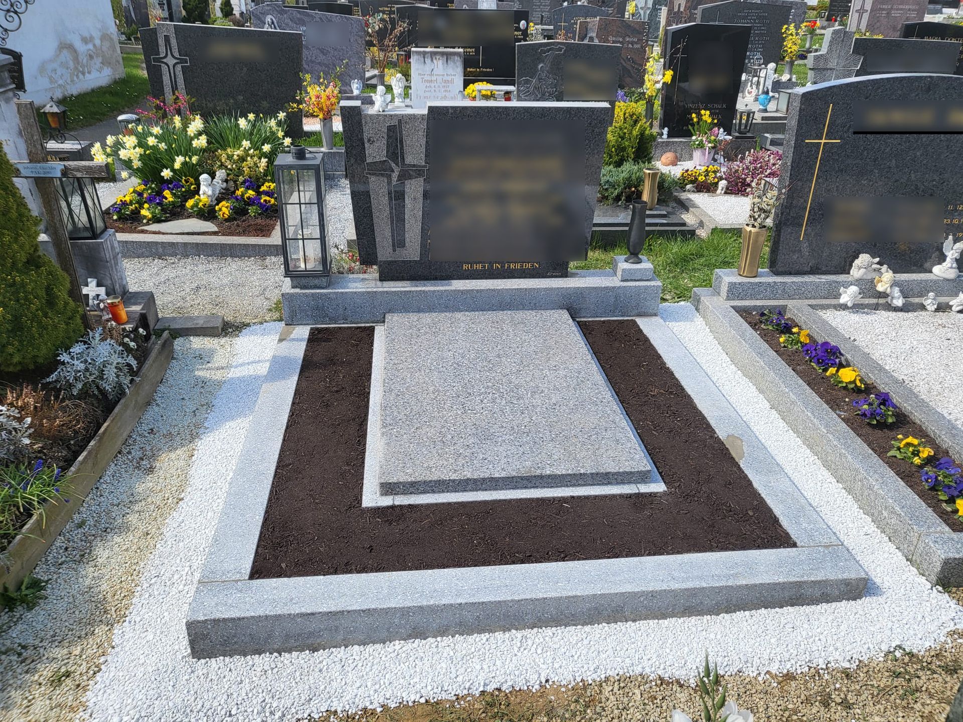 Grab mit grauem Steinsockel und Gedenktafel, umgeben von Blumen auf einem Friedhof.