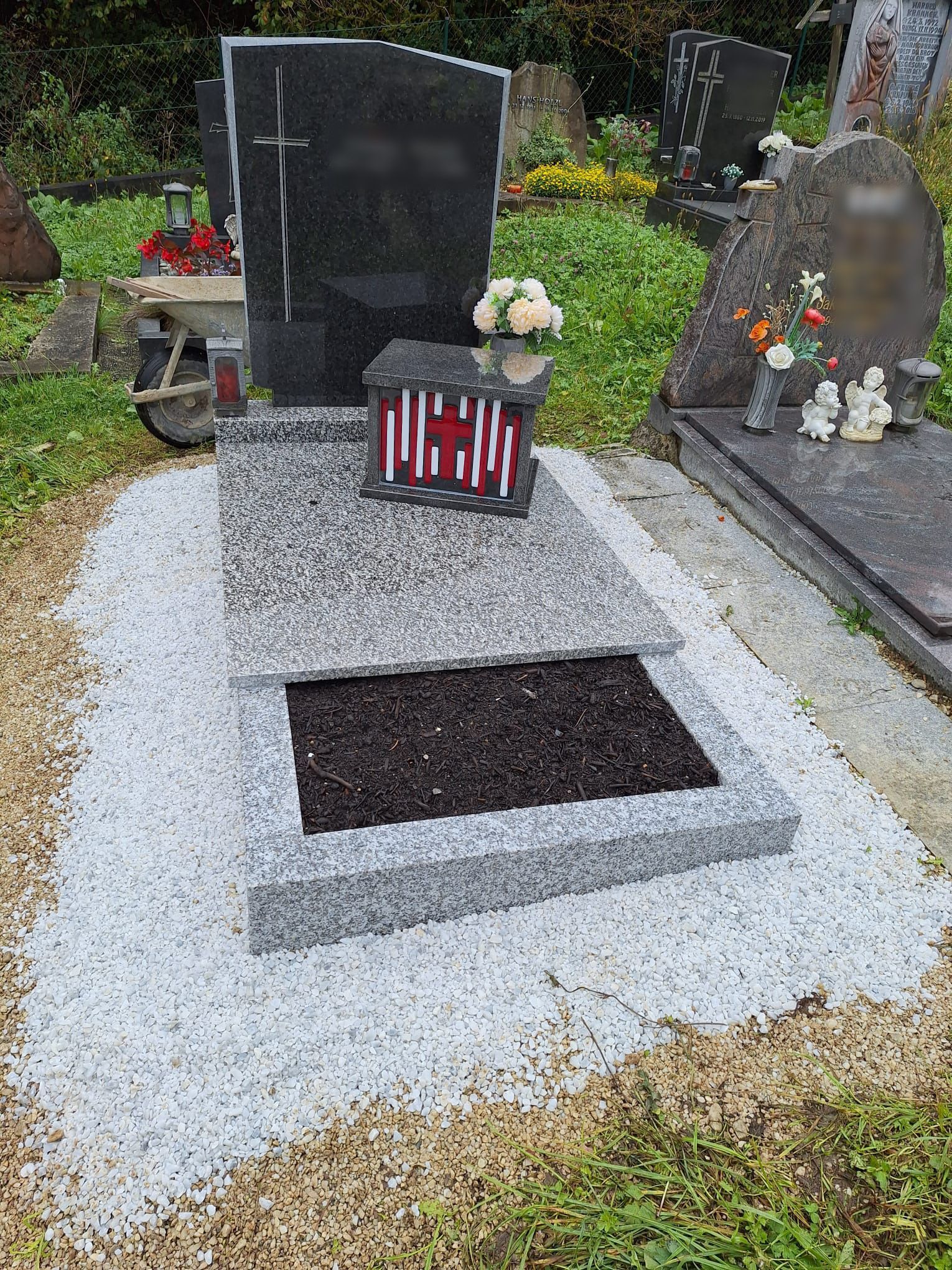 Mit Kies bedecktes Grab mit schwarzem Grabstein, roten Blumen und einer kleinen Gedenklaterne auf einem Friedhof