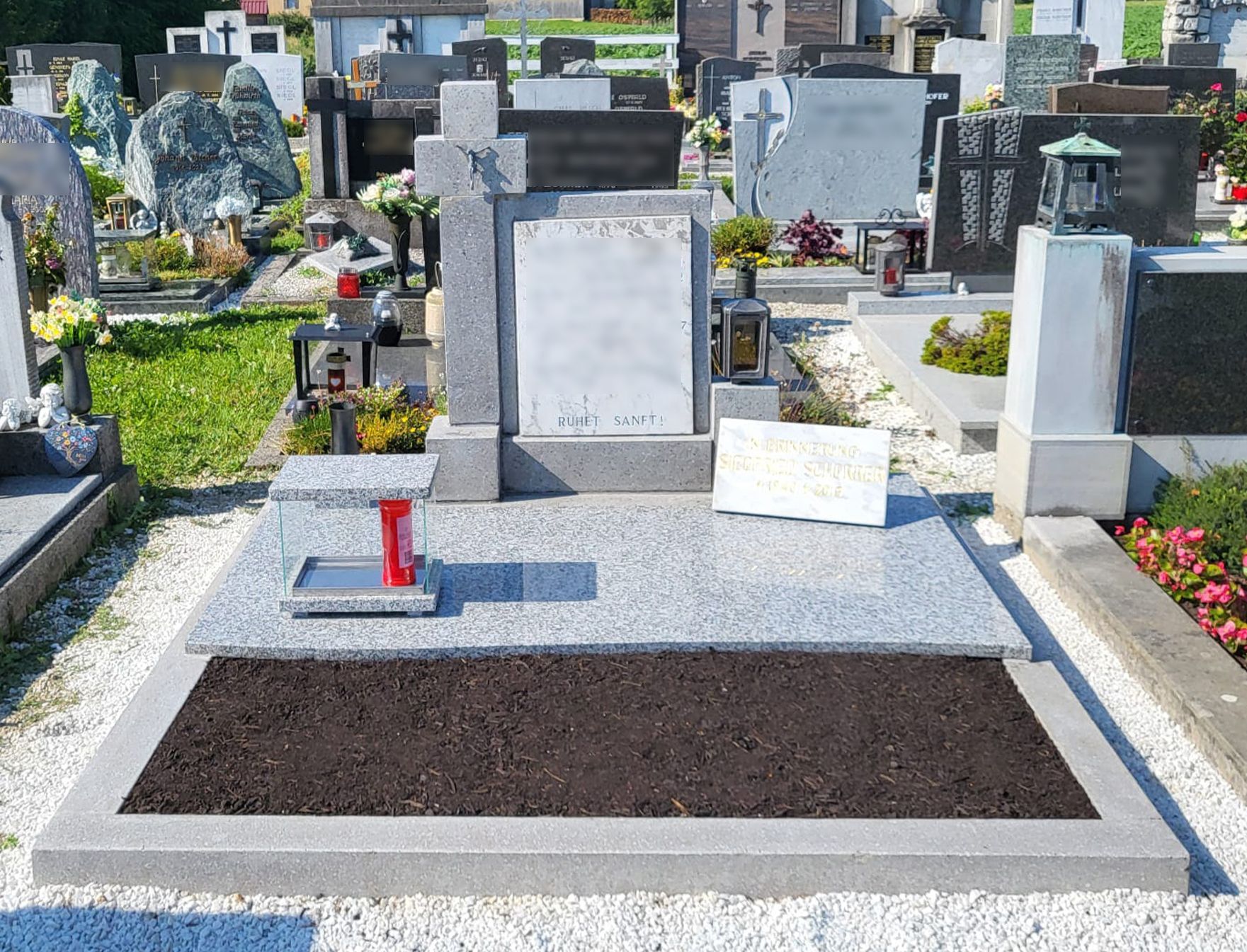 Grabstein mit frischen Blumen und einer leeren Gedenktafel auf einem Friedhof, umgeben von anderen Grabdenkmälern.