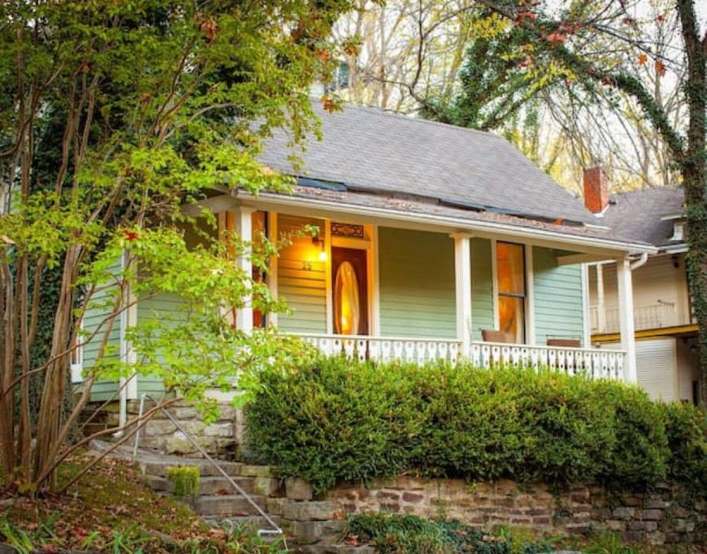 White house with a lit porch, surrounded by trees and shrubs on a stone retaining wall.