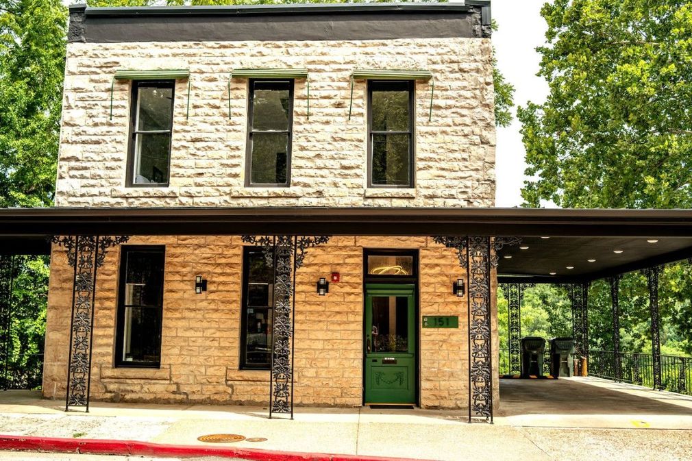 Historic stone building with green doors and covered porch beside a tree-lined street