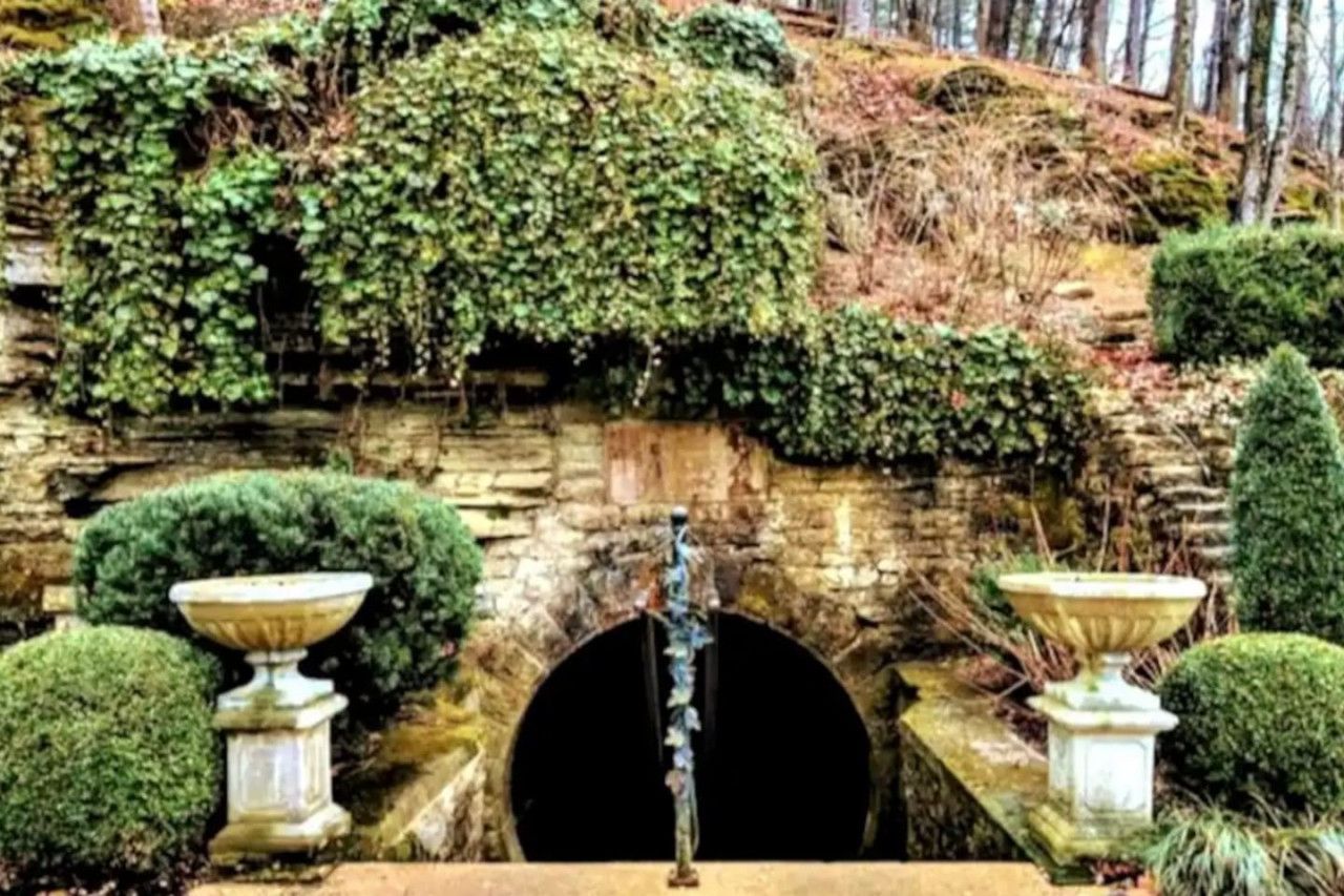 Stone grotto fountain with arched opening, flowing water, and trimmed topiary in a garden setting