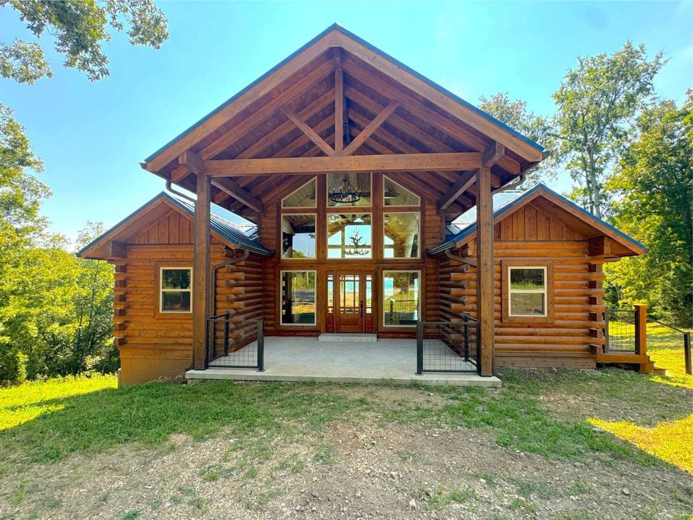 Log cabin-style house with a timber porch and large windows, surrounded by trees and grass