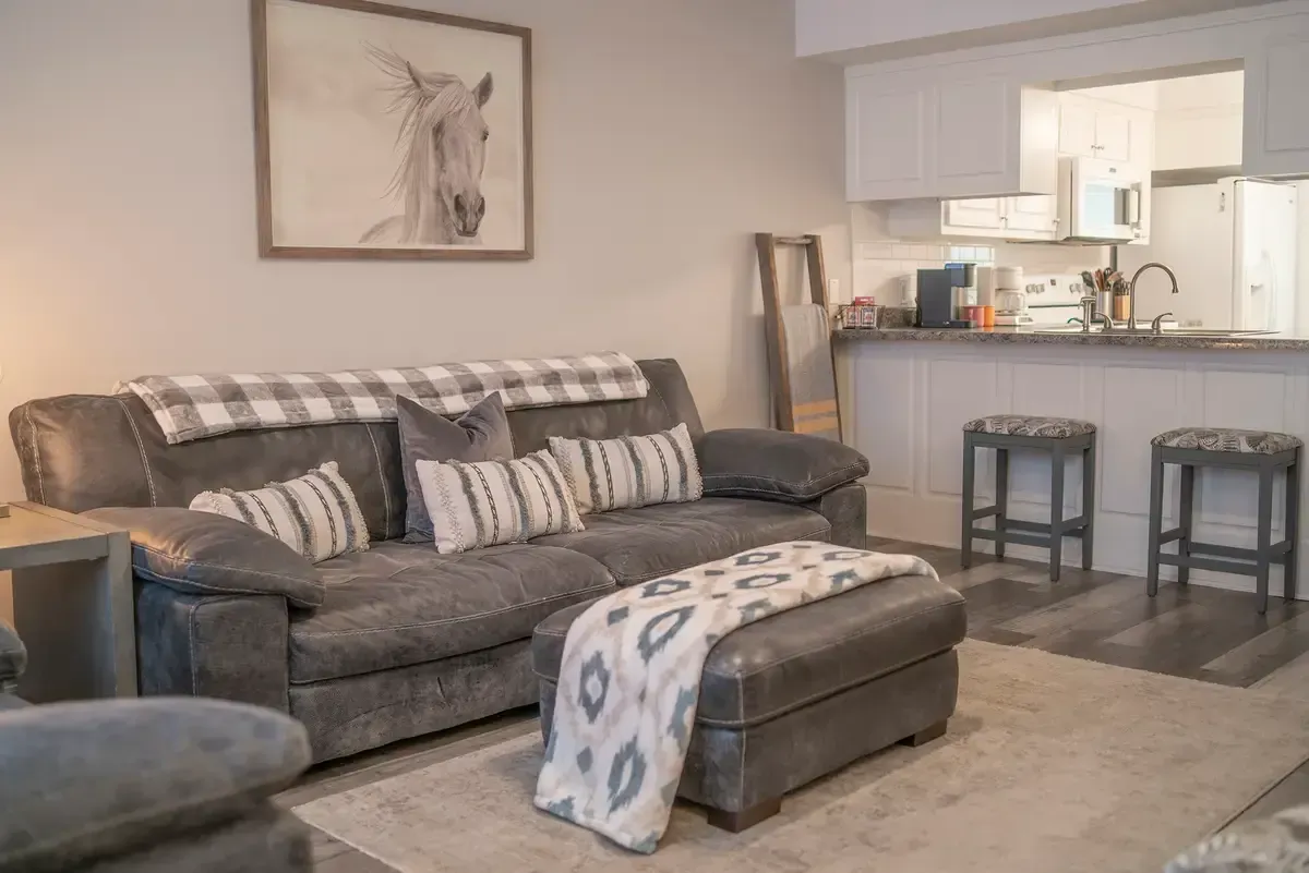 Cozy living room with gray sofa, ottoman, framed art, and open kitchen with bar stools