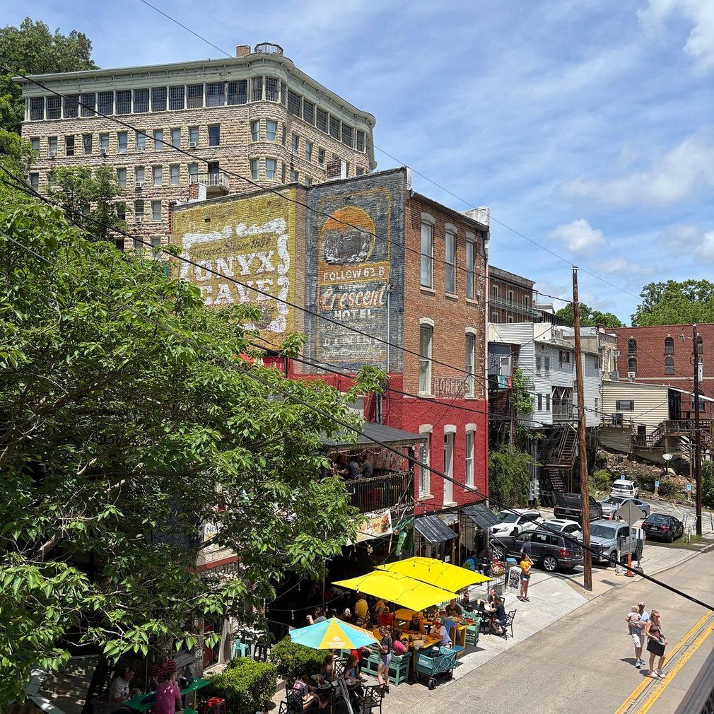 Busy city street with outdoor cafés, colorful umbrellas, brick buildings, and a large mural on a sunny day