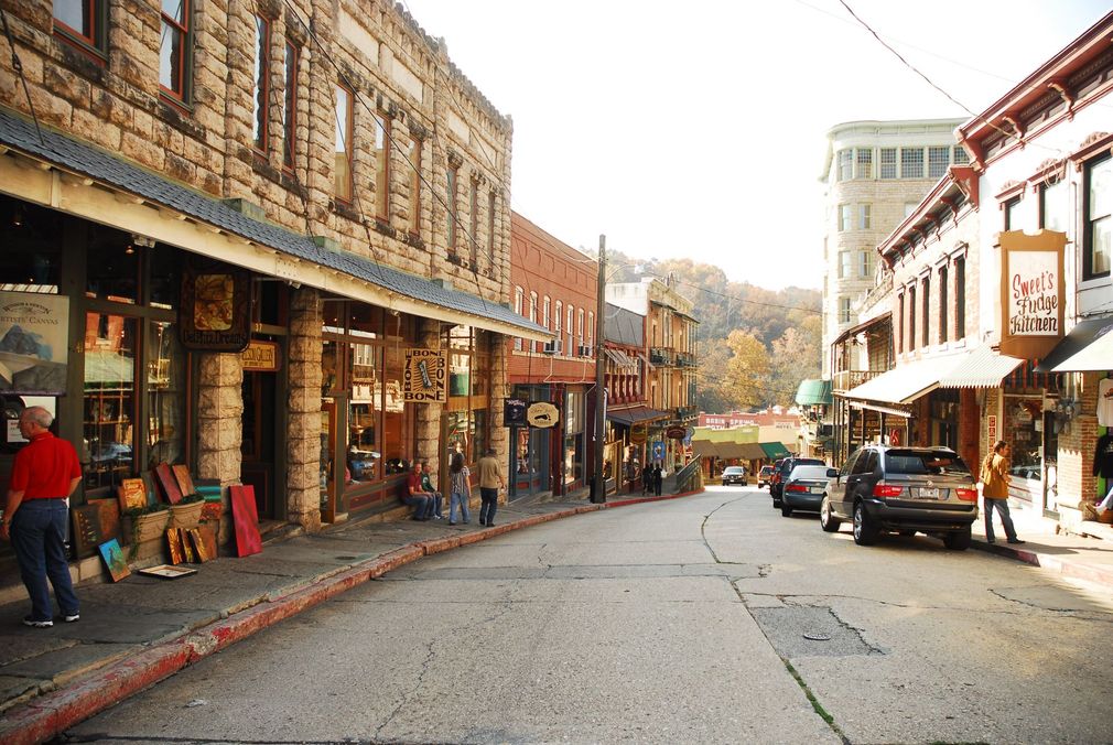 Historic downtown street with shops, parked cars, and a few pedestrians in warm sunlight