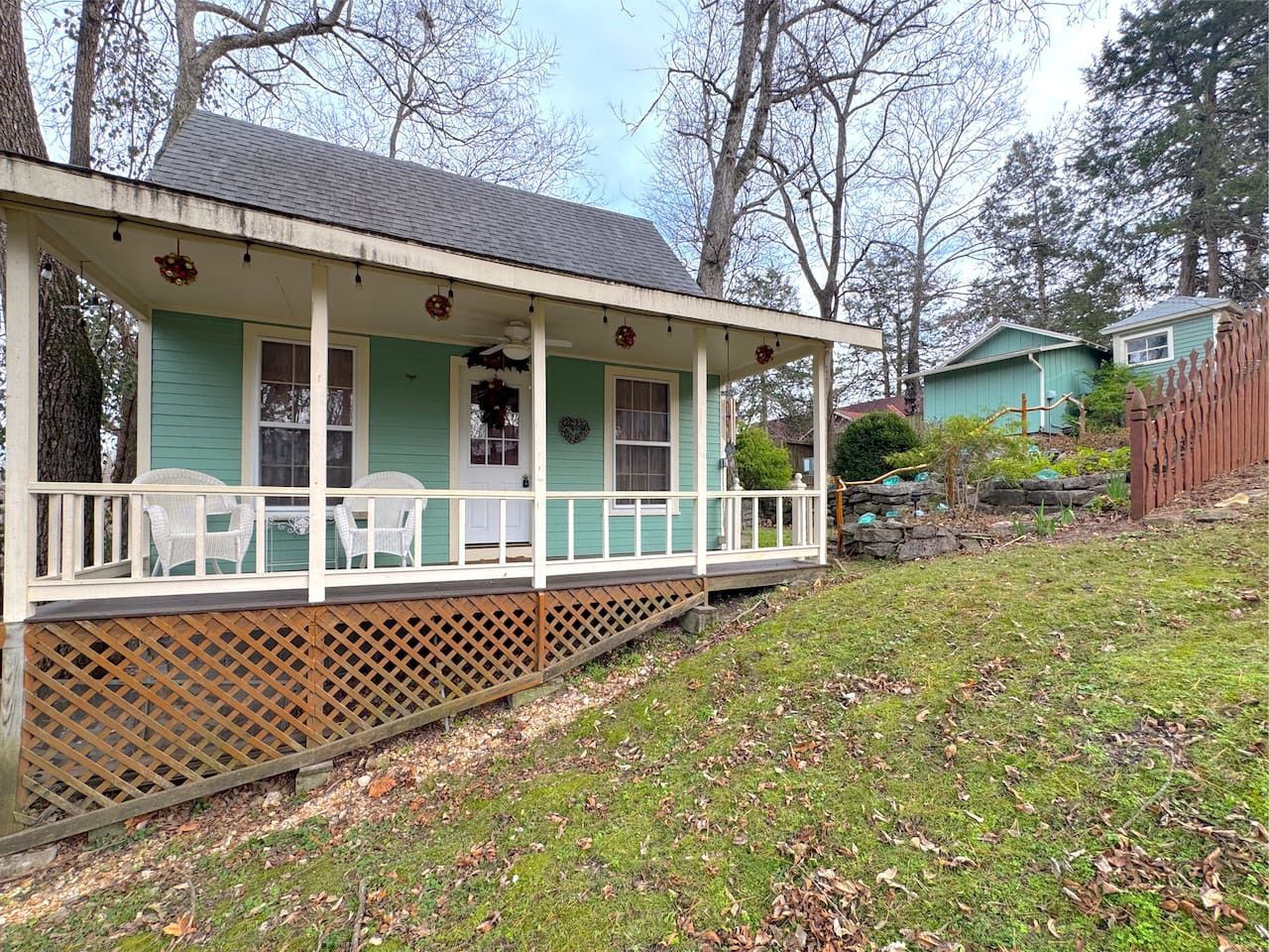 Small turquoise house with white porch on a grassy hillside, surrounded by trees and a fence