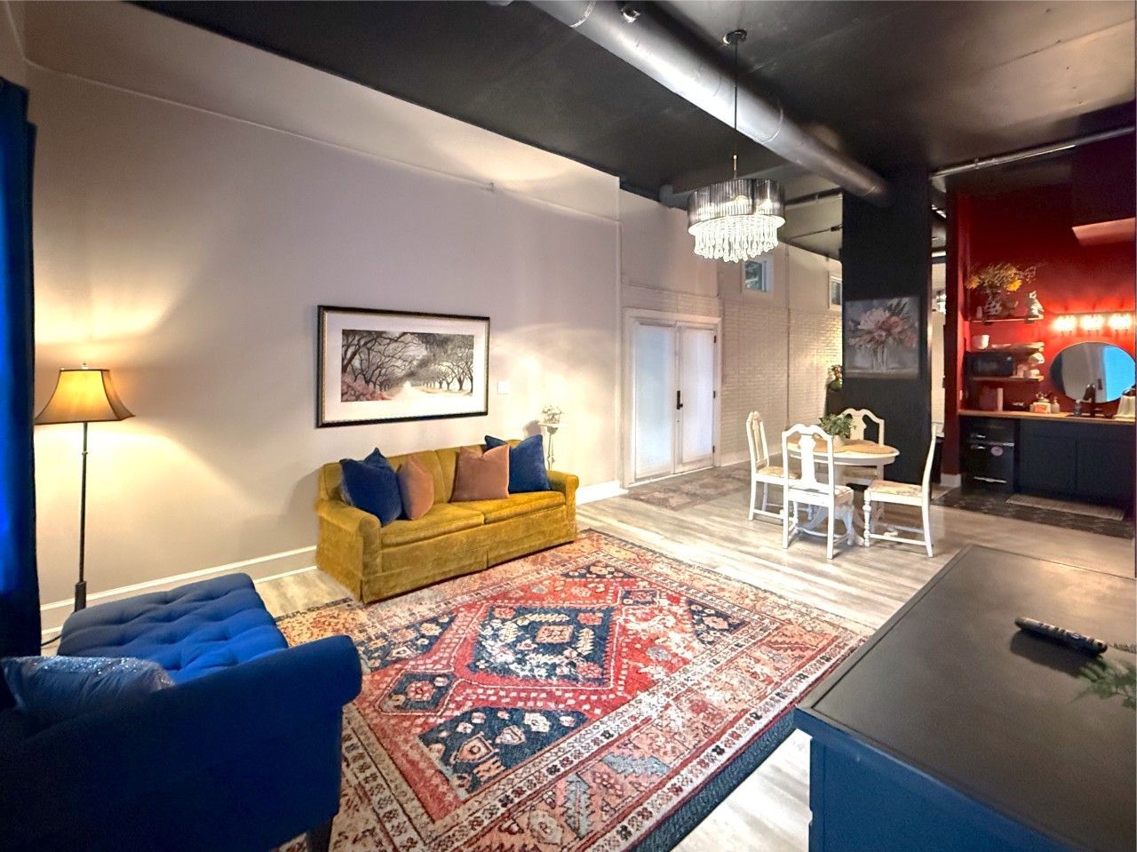 Modern living room with blue and mustard sofas, patterned rug, chandelier, and dining table by red-lit kitchen