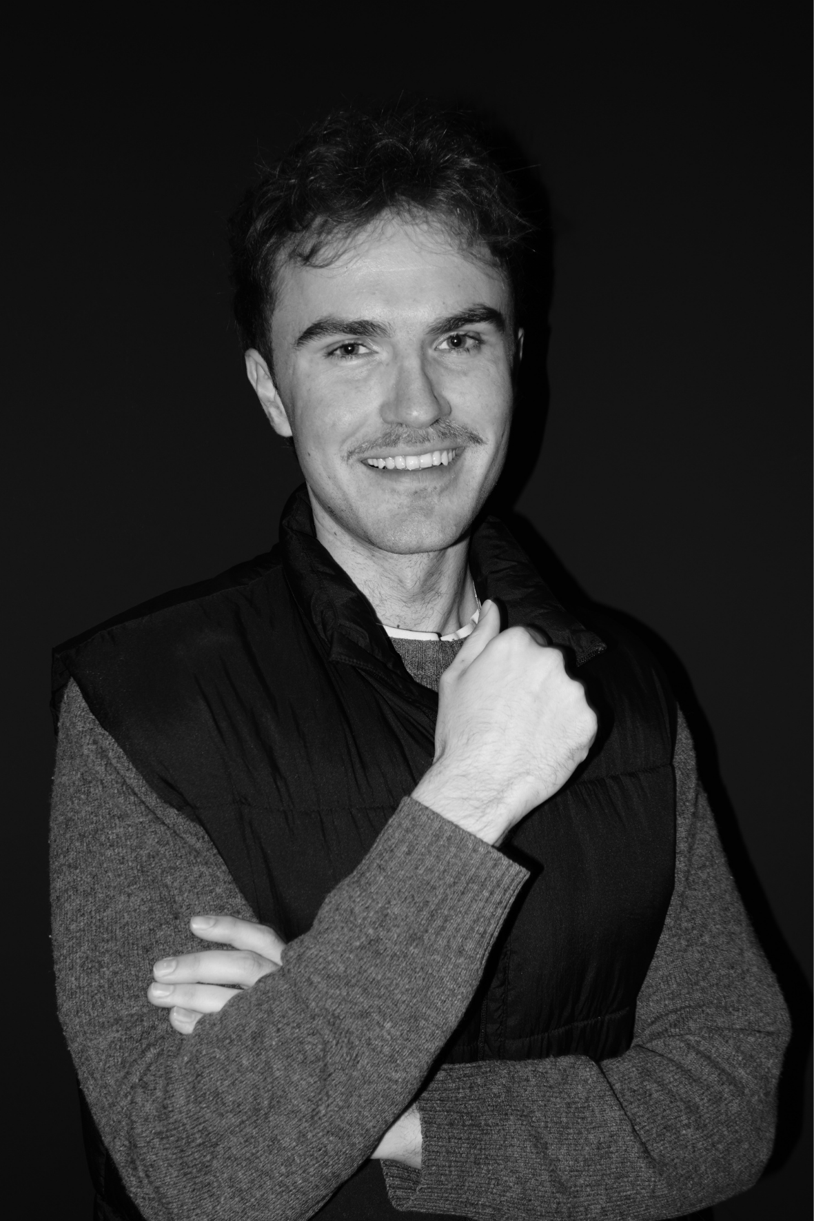 Man smiling in a dark vest over a gray sweater, standing with arms crossed against a dark background