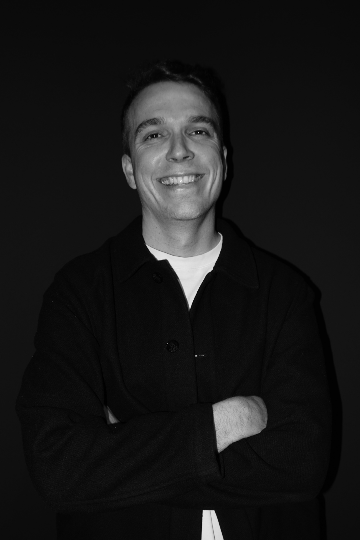 Smiling person in a dark shirt, standing with arms crossed against a black background