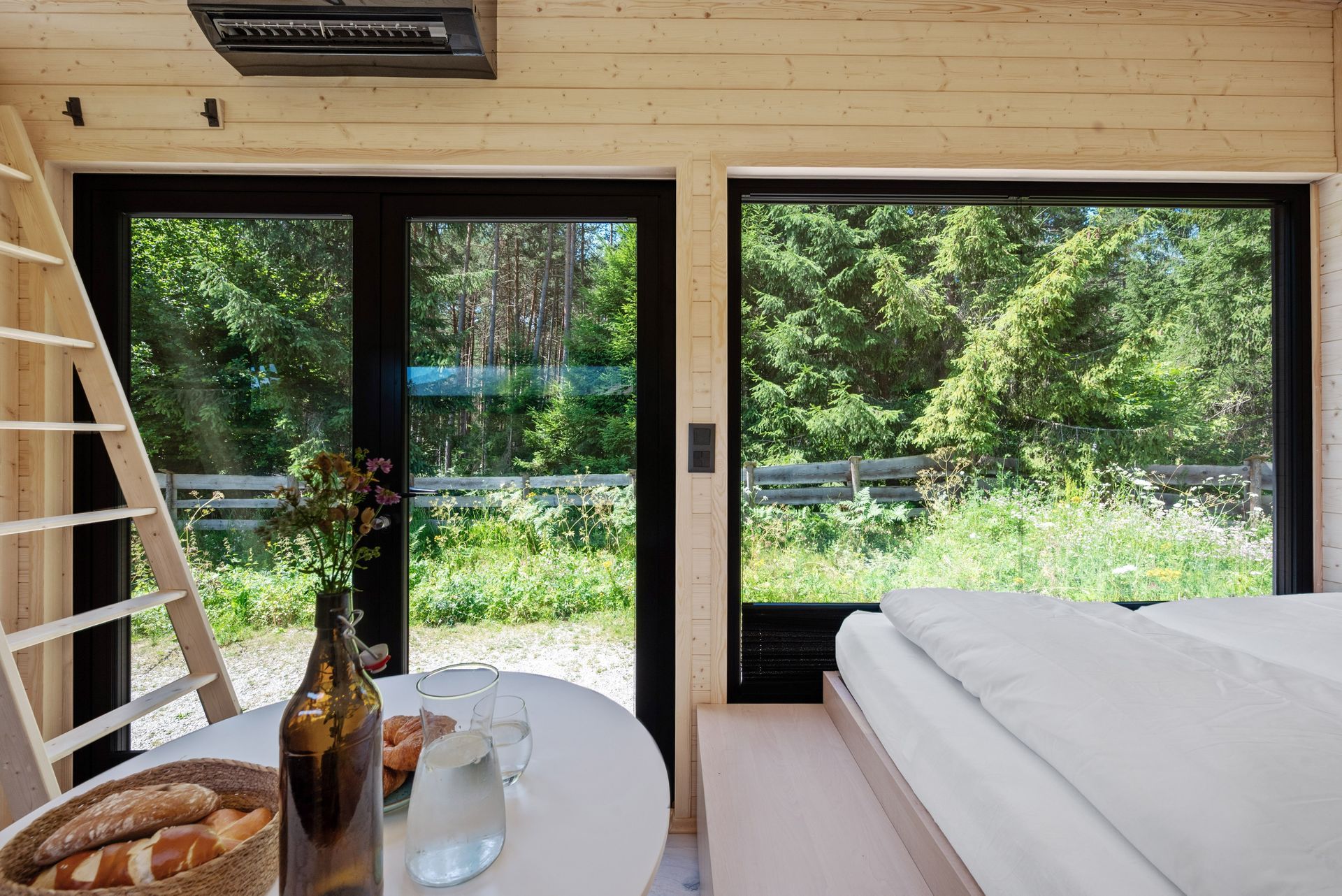 Small dark wooden structure in a gravel clearing, surrounded by tall green trees and wildflowers.