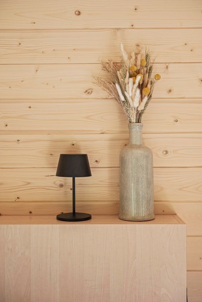 Minimalist wooden shelf with a small black lamp and a tall vase of dried flowers against a light wood wall