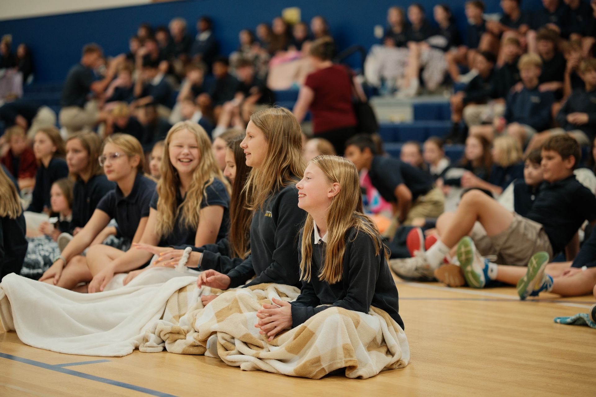 Students in blue uniforms sit on floor of gymnasium, watching event. Some smile.