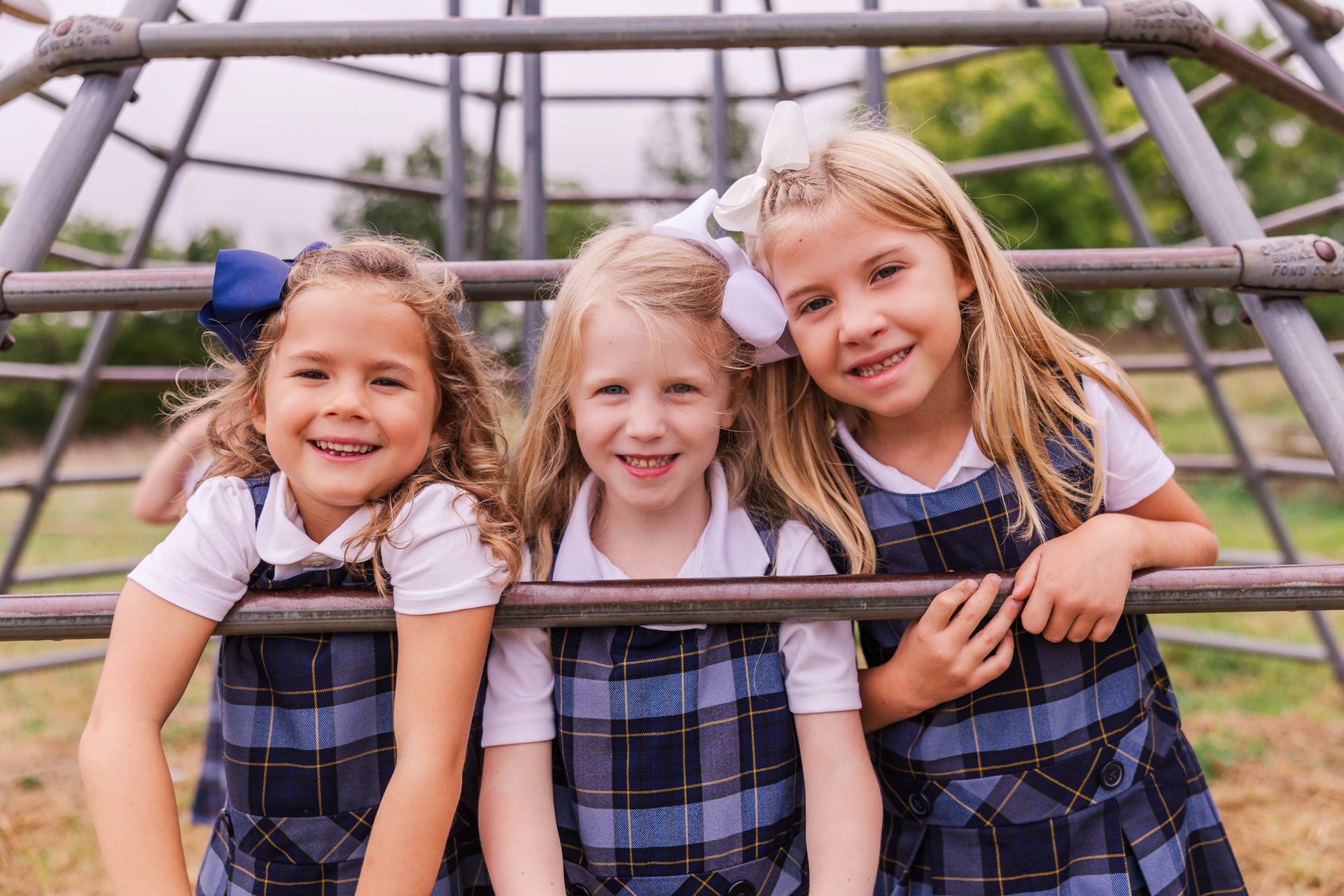 Three girls in plaid jumpers smile at camera, leaning on a playground structure outdoors.