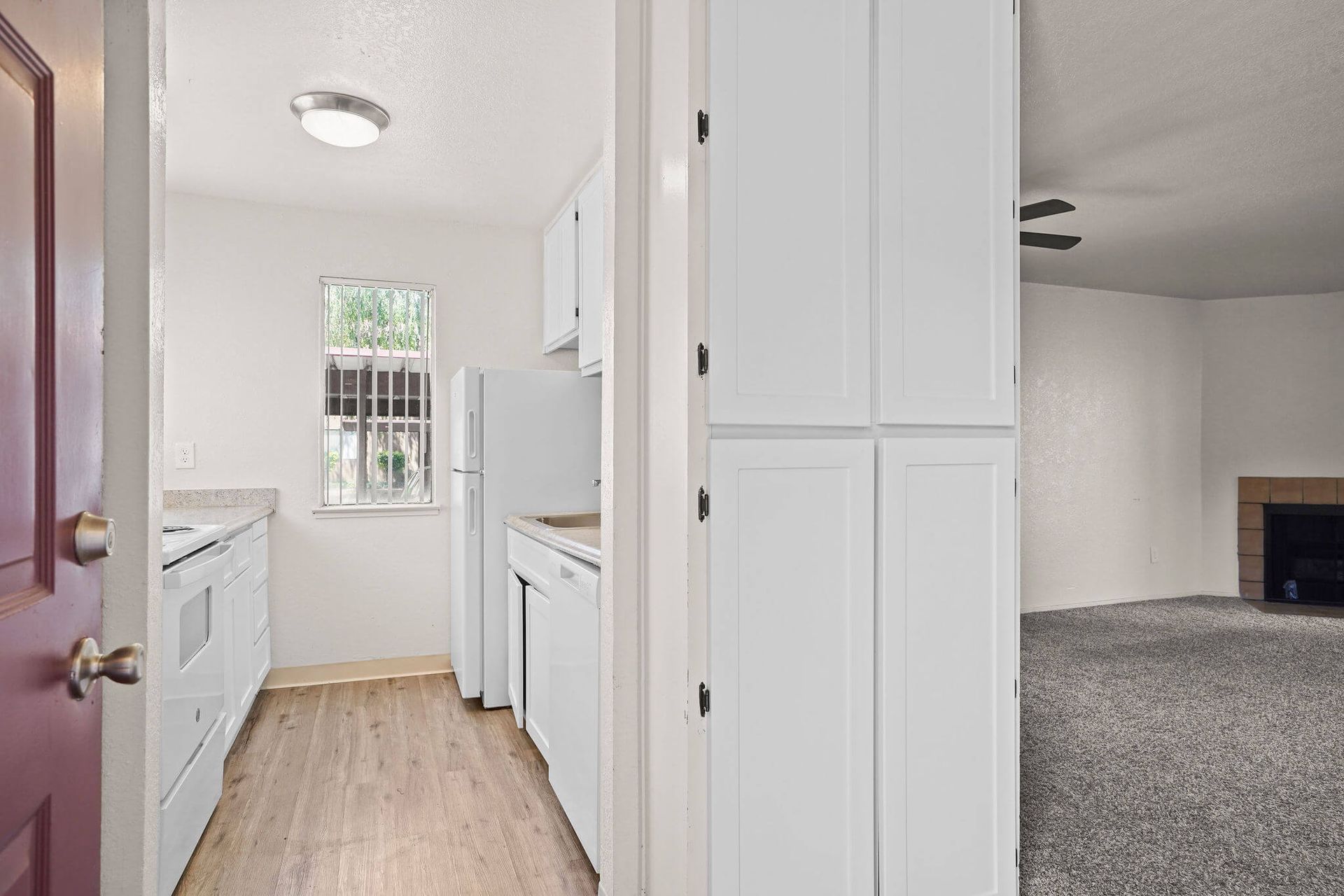 Bright narrow kitchen hallway with white cabinets, white appliances, and a window at the end, opening to a carpeted room.