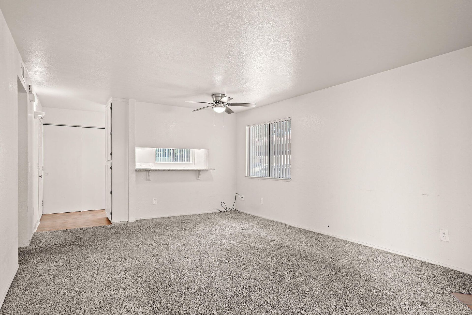 Empty white living room with gray carpet, ceiling fan, window, and wall shelf.