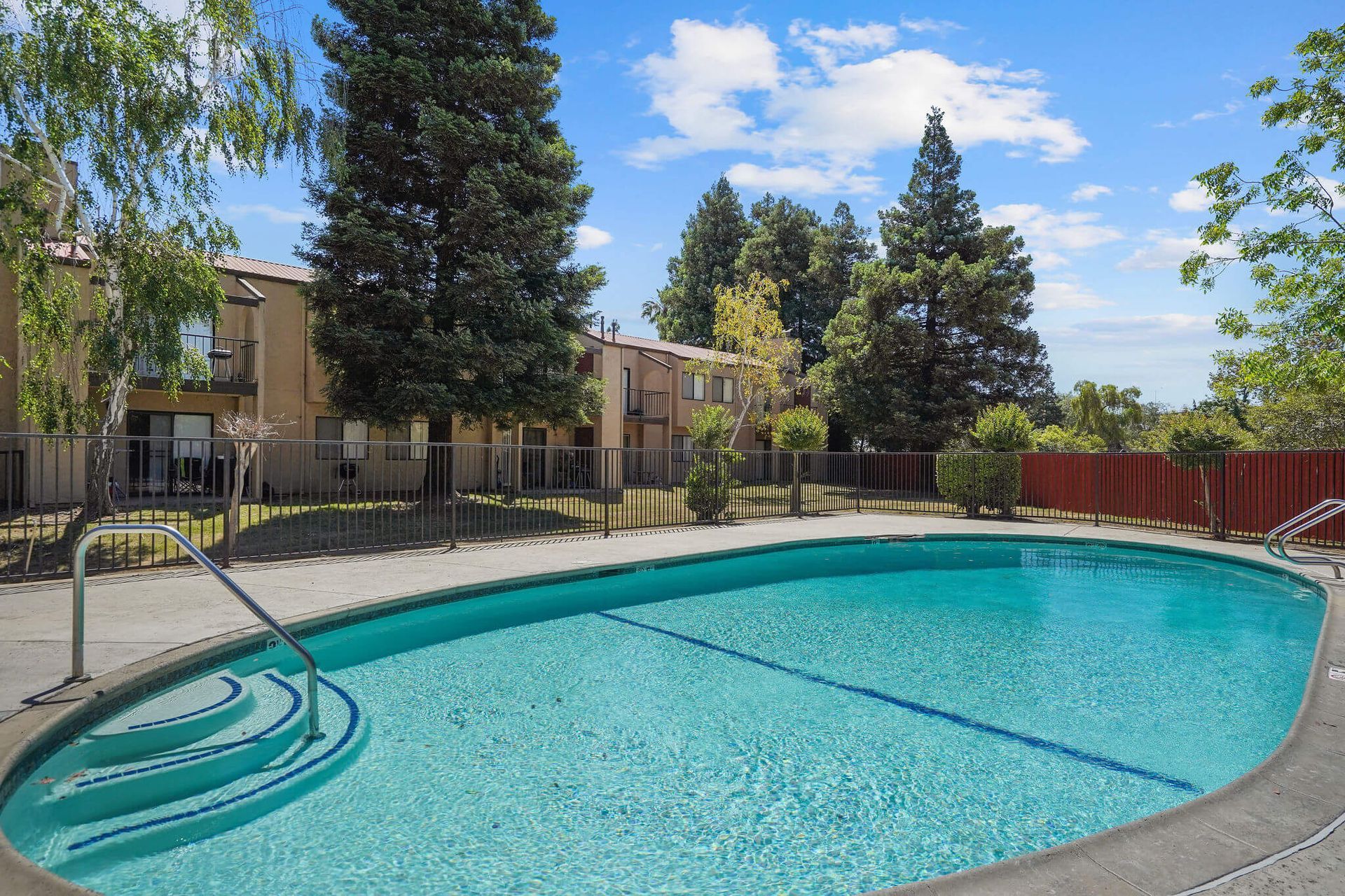 Outdoor swimming pool with clear blue water, metal handrail, and apartment buildings and trees in the background