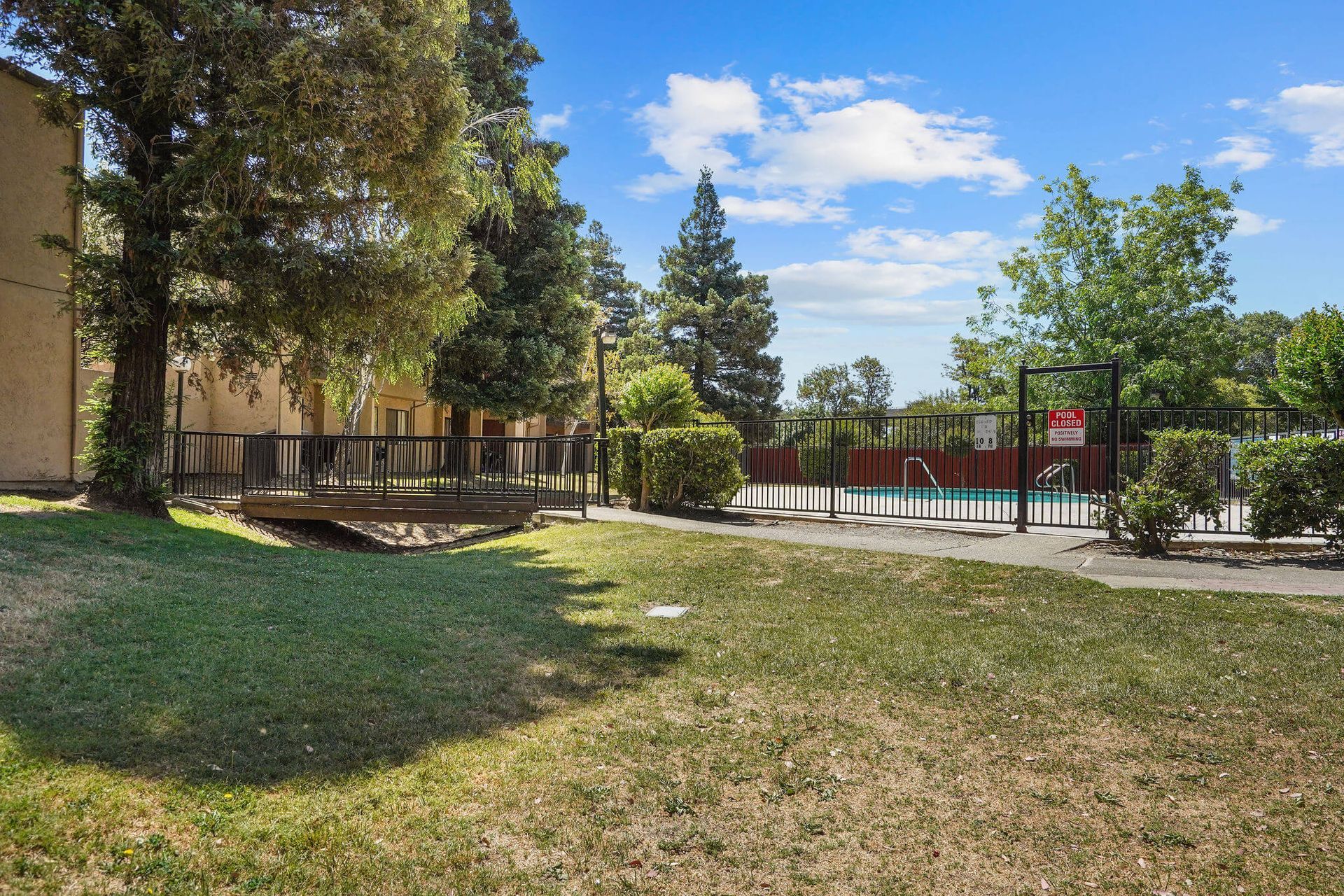 Sunny courtyard with trees, grass, and a fenced playground beside a building