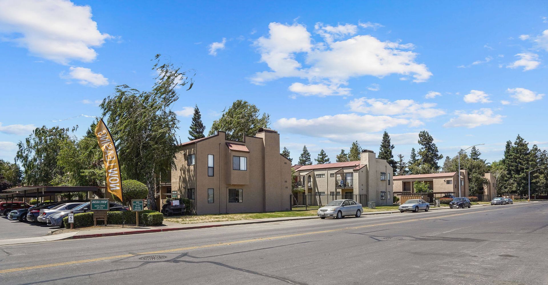 Apartment complex parking lot with beige buildings, trees, and a blue sky with clouds
