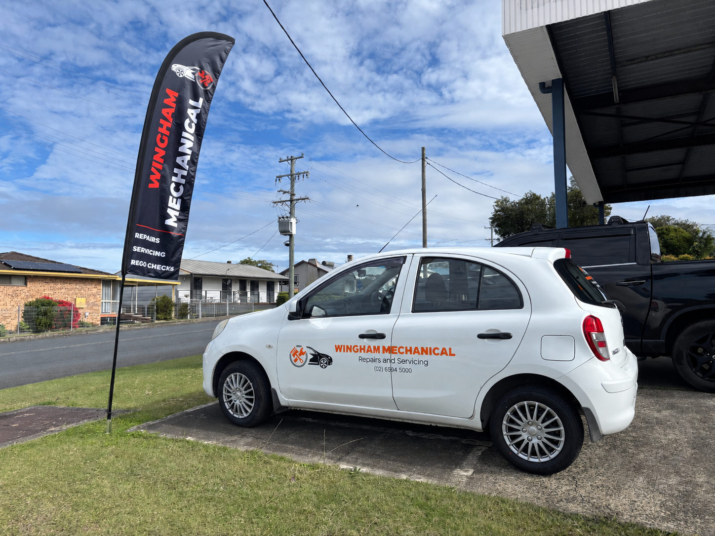 White Service Car Parked Outside a Building Beside a Black Promotional Flag on a Sunny Day — Wingham Mechanical in Wingham, NSW