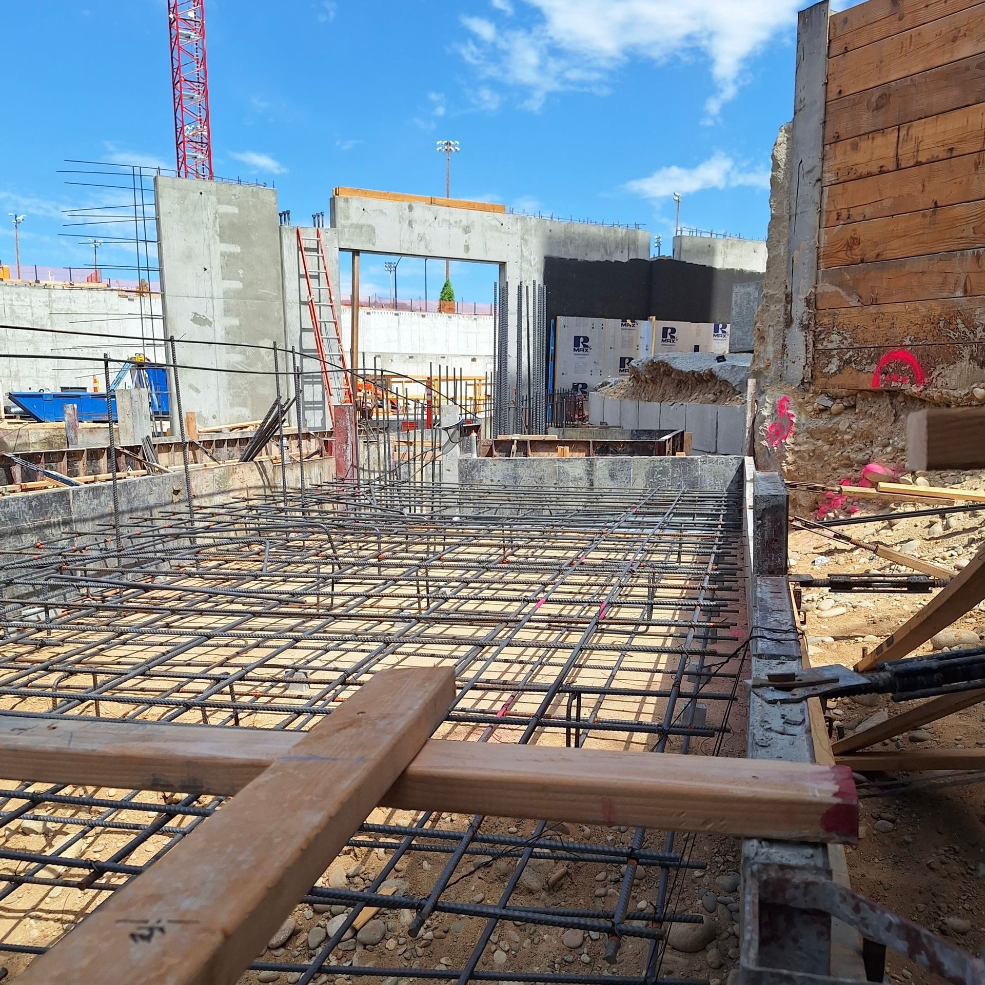 Construction site with rebar grid, concrete walls, and crane under a blue sky.