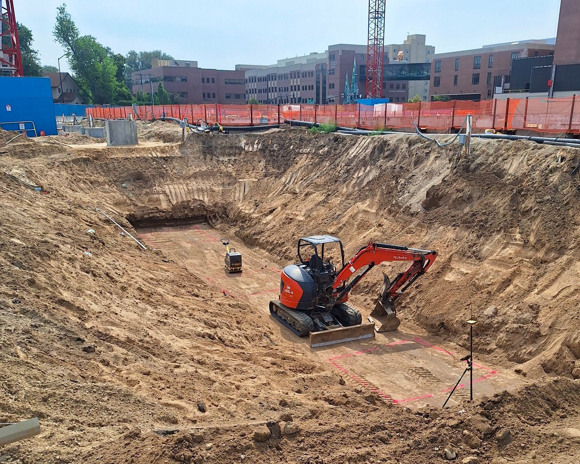 Construction site with excavator digging in a large pit. Buildings and fencing visible in background.