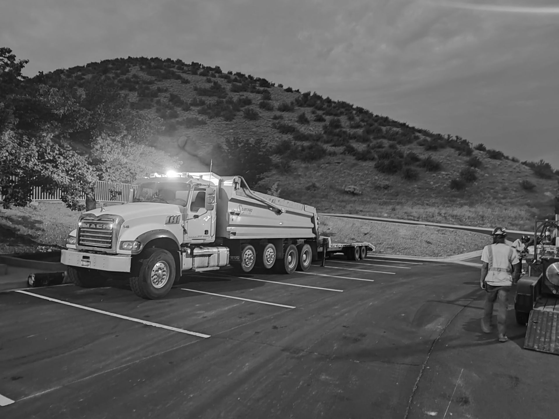 Large dump truck on road at dusk, lights on, with worker nearby and hillside in background.