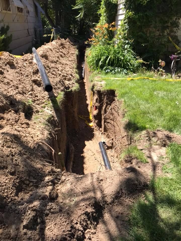Trench dug in a yard with black pipes, dirt, and grass, near a house.
