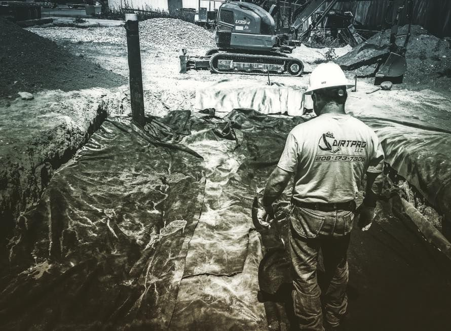 Construction worker with tool in hand, surveying trench; excavator in background.