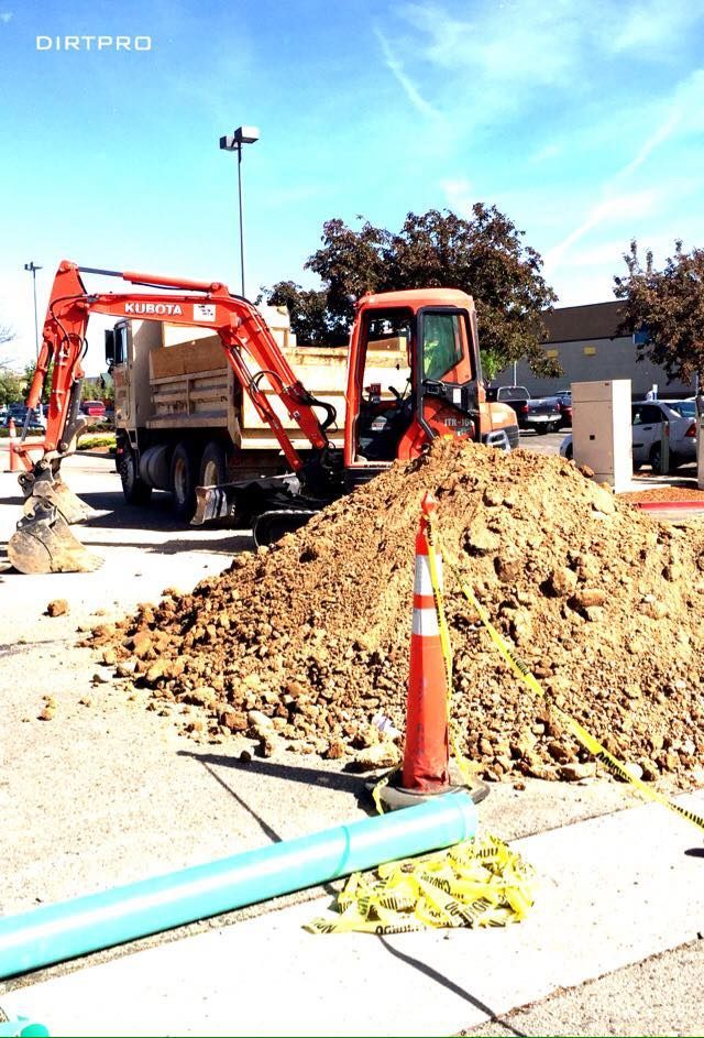 Excavator loading dirt into dump truck in a construction site with caution cones and blue pipe.