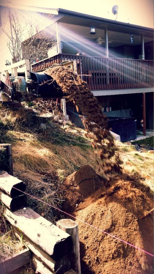 A backhoe dumping dirt from a hill near a house with a porch and wooden railing.