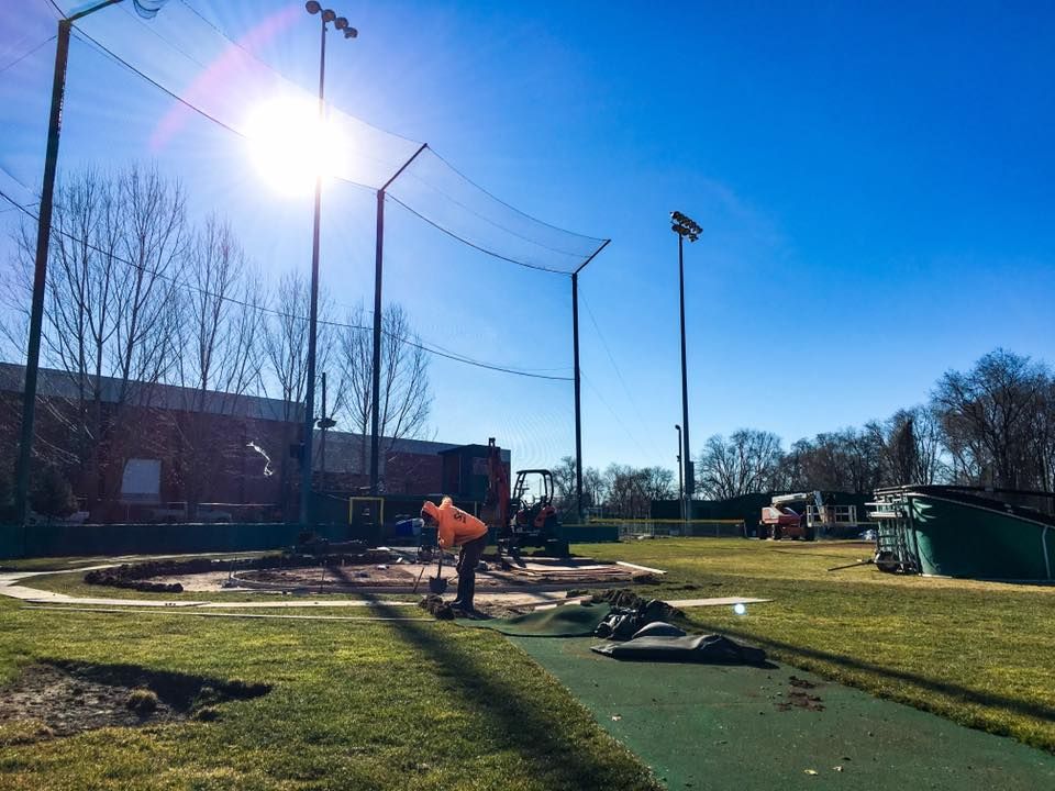 Construction at baseball field: workers, protective netting, bright sun.