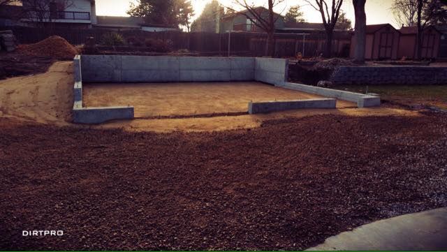 Concrete foundation walls being constructed; dirt ground and gravel surround.