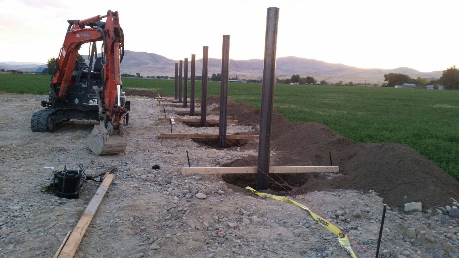 Mini excavator installing fence posts in a field, with mountains in the background.