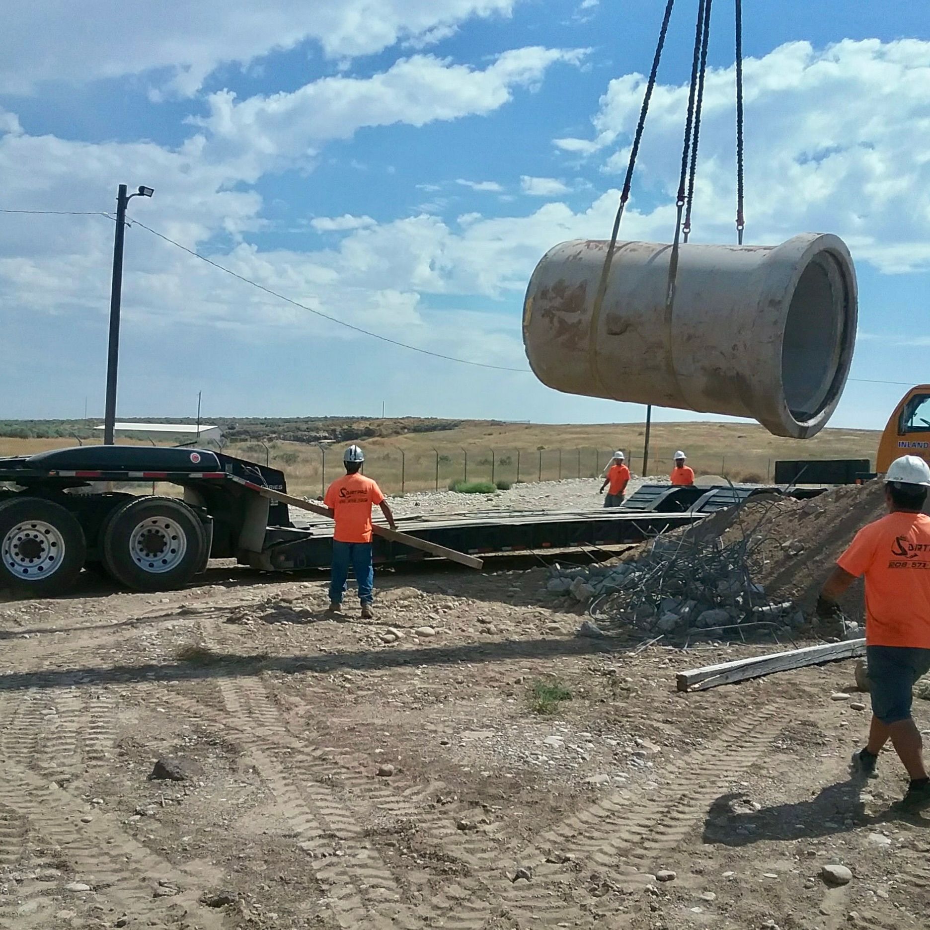 Construction workers lifting a large concrete pipe with a crane. Setting is a dirt field under a blue sky.